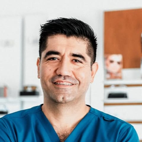 El Dr. Juan Antonio Liera García, sonriendo, con uniforme médico azul y el aparador de armazones de su óptica detrás