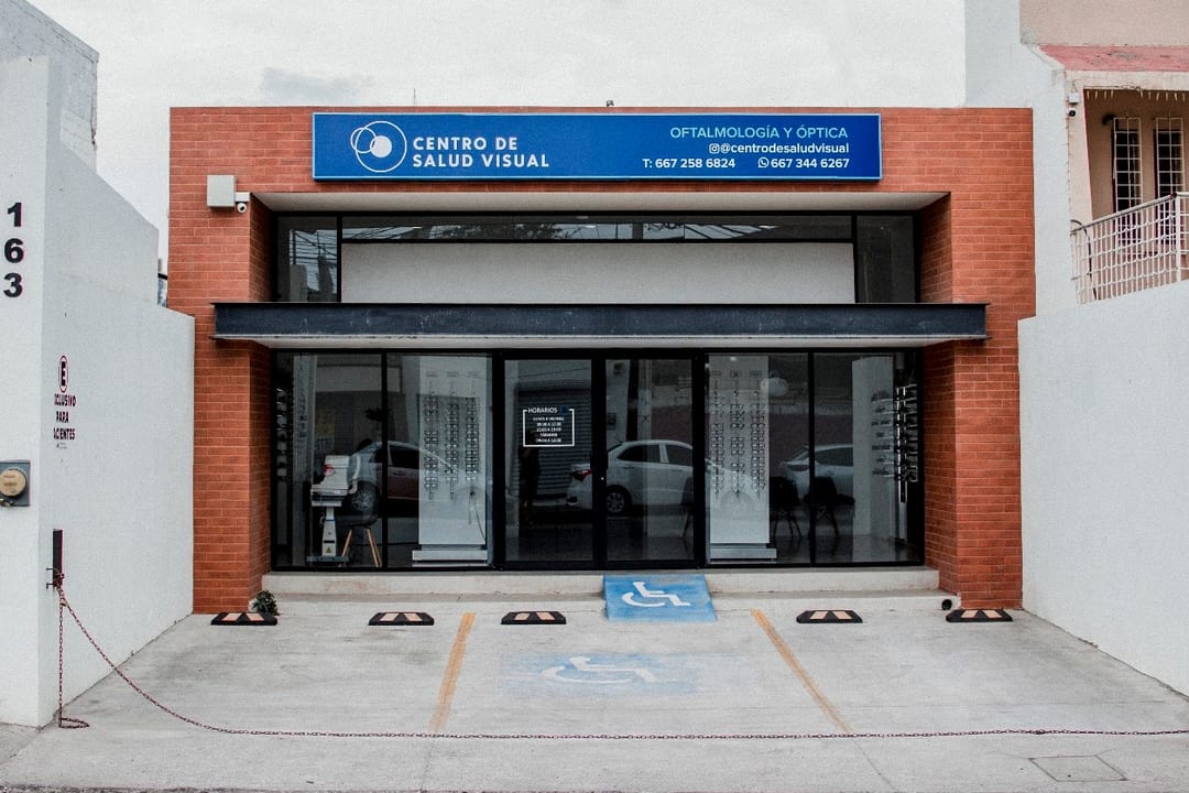 El local del Centro de Salud Visual visto de frente: fachada de ladrillo con letrero azul, aparador de cristal a la calle y, enfrente, un estacionamiento con un cajón marcado para personas con discapacidad
